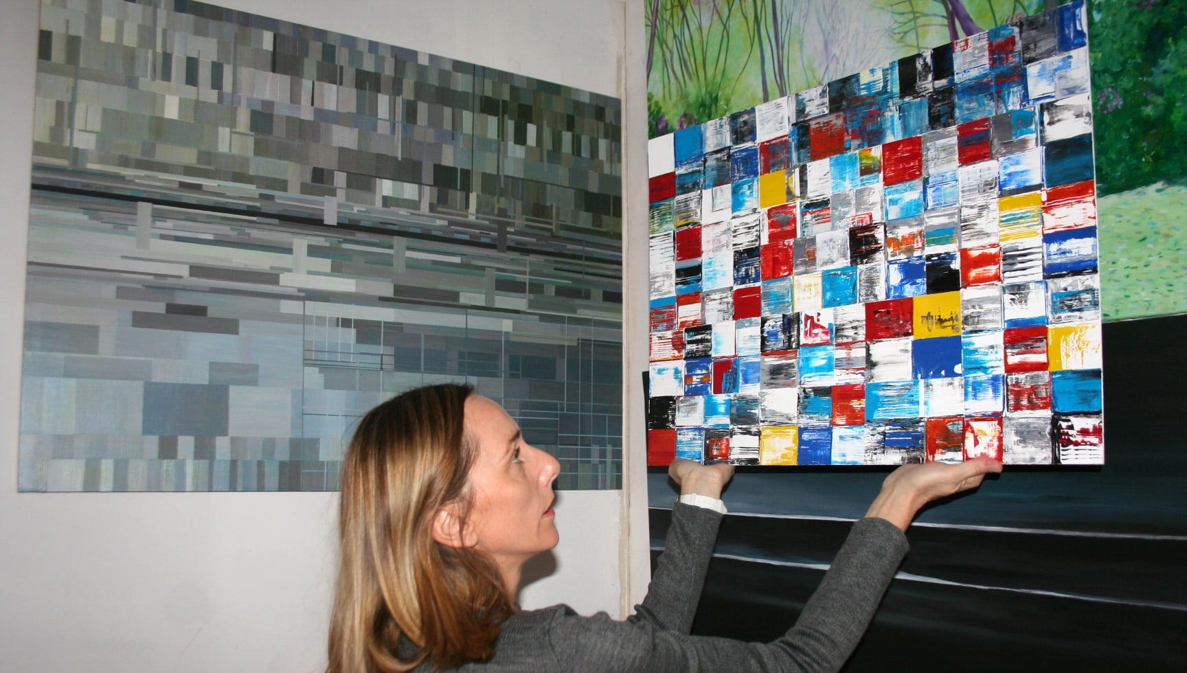 Nadine Hardy dans son atelier, tenant à bout de bras une composition de carrés rouges, bleus et jaunes devant deux grandes toiles